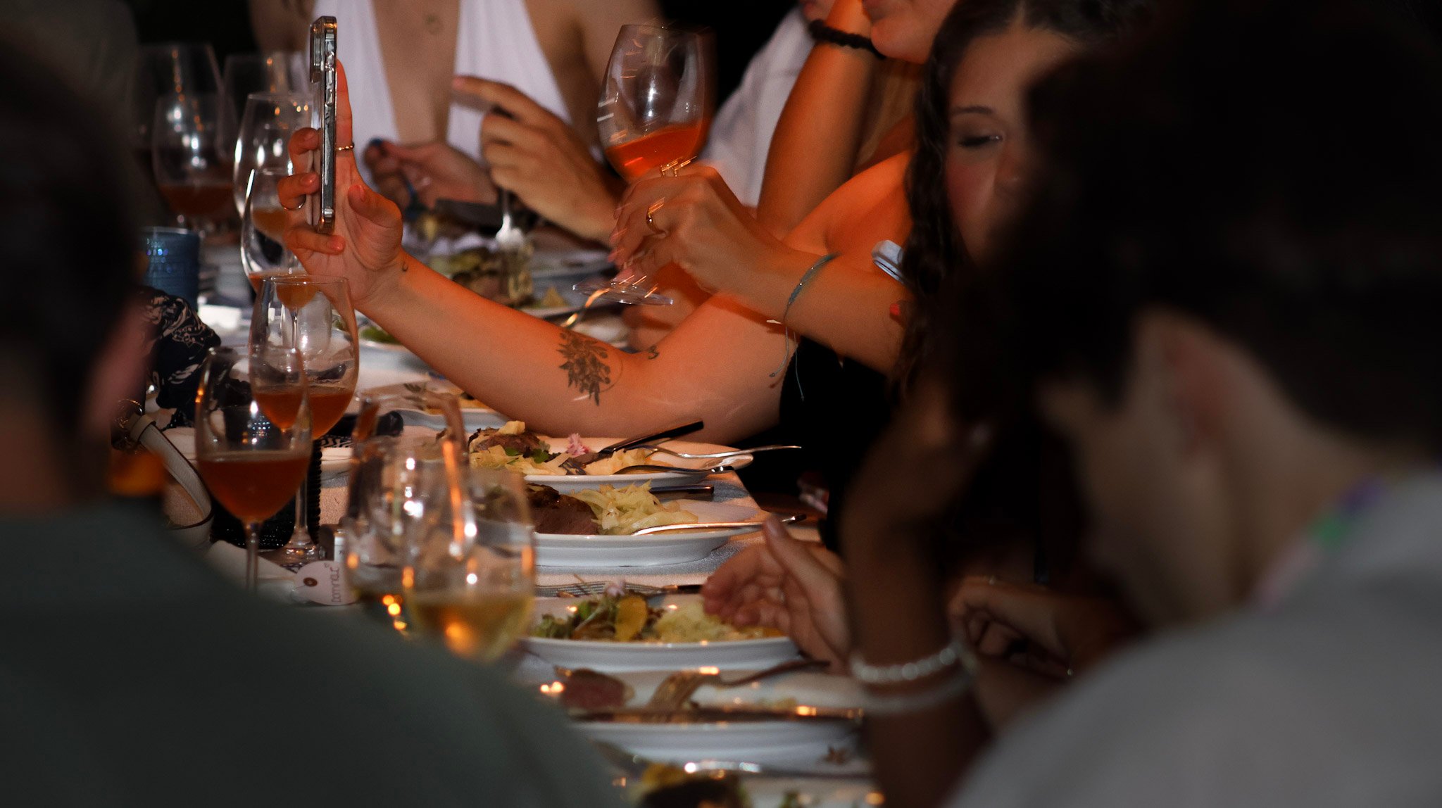 Diners gather around a candlelit table with food, wine glasses, and a person holding a smartphone.