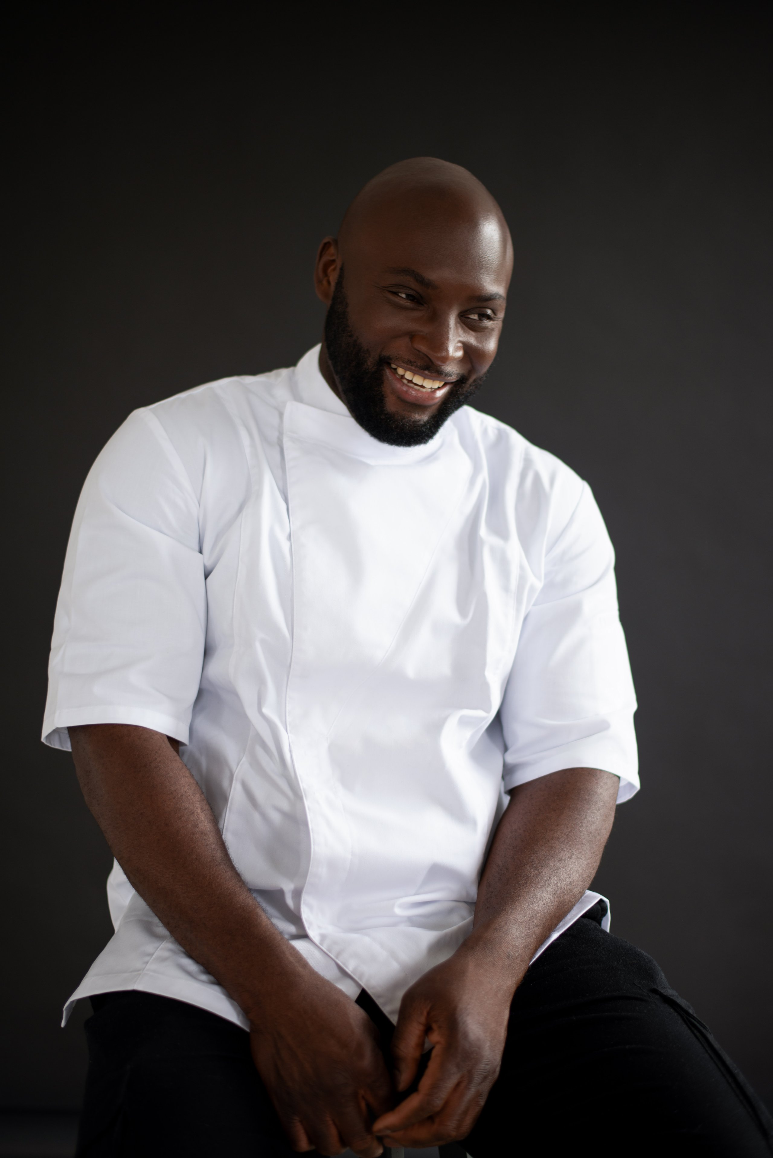 A smiling Black chef with a shaved head and beard in a white short-sleeved jacket against a dark background.