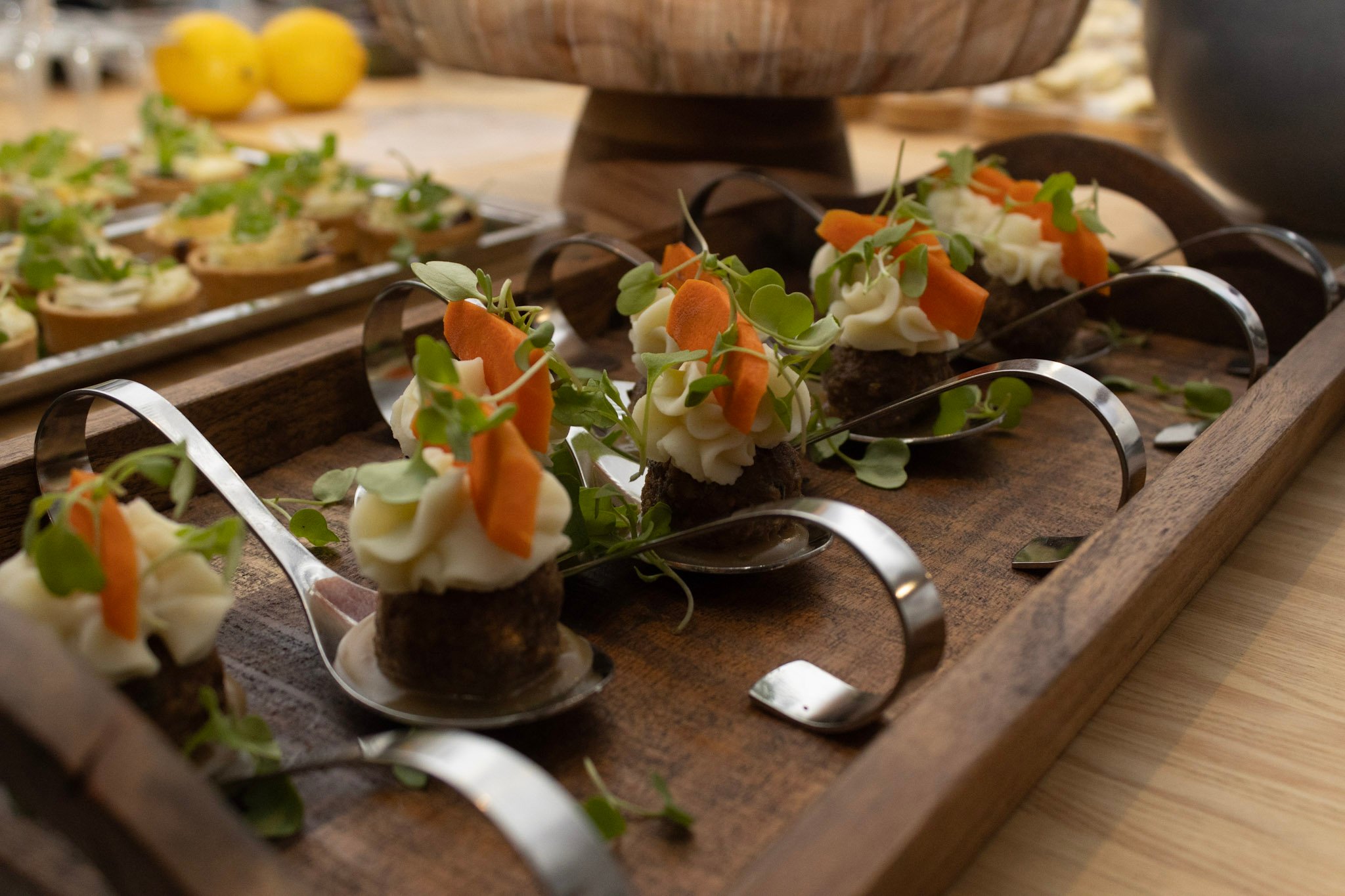 Bite-sized appetizers with white cream, orange vegetable slices, and microgreens arranged in silver spoons on wooden trays.