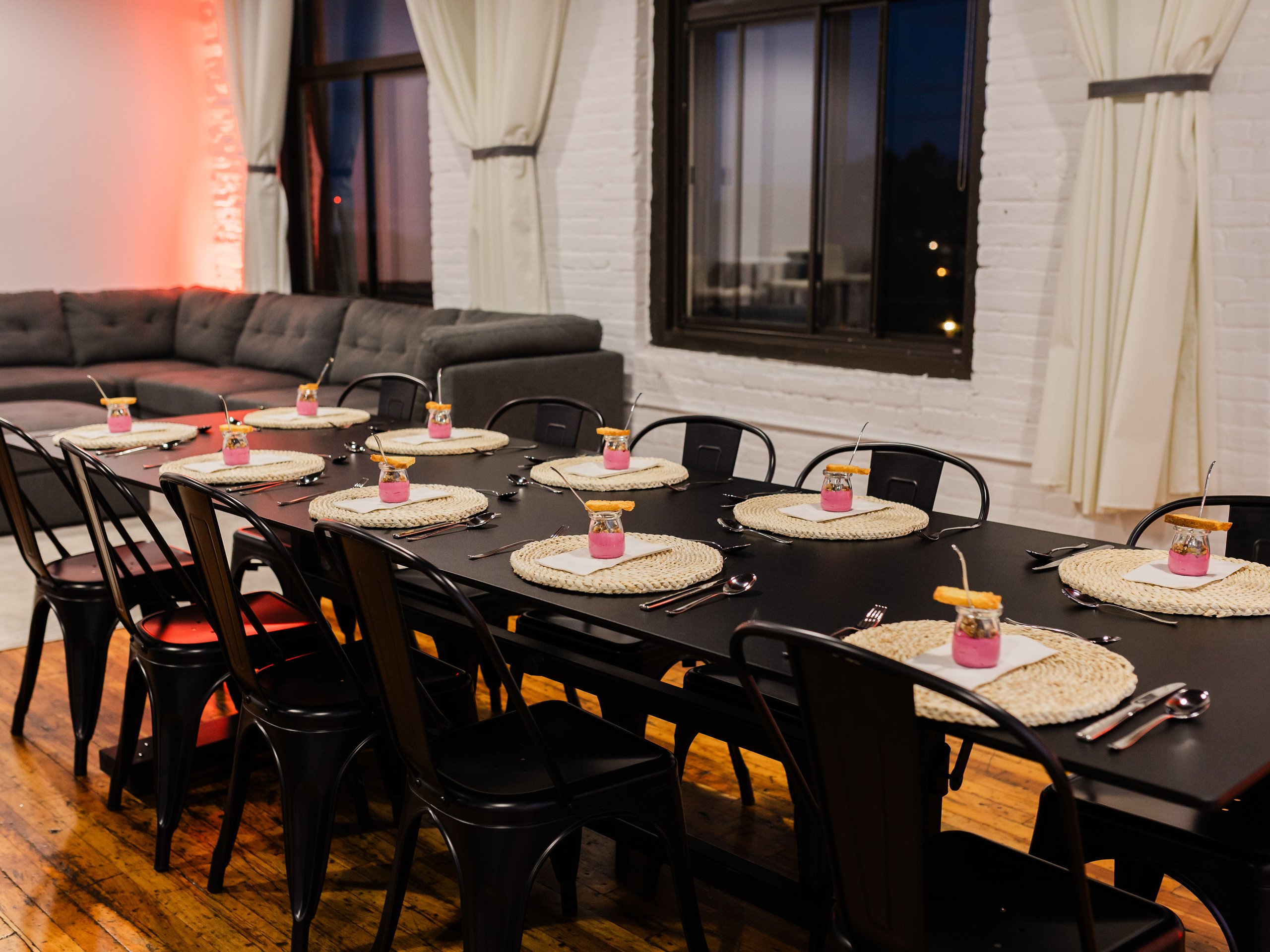 Warmly lit restaurant dining room with set black tables, pink drinks, black chairs, sofa, windows, and white brick walls.