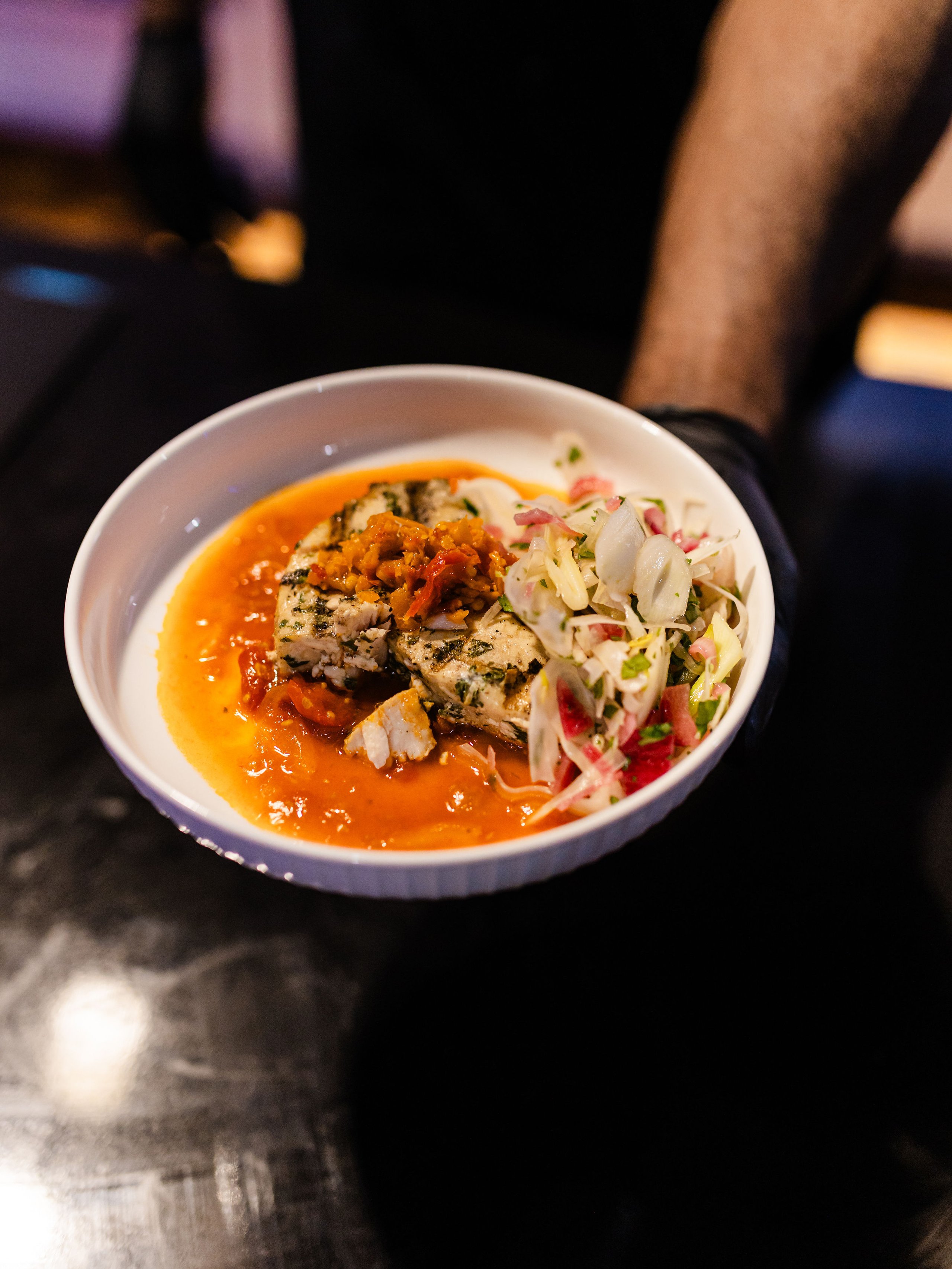 White bowl of sauced grilled food, orange grains, and shredded salad against a dark blurred background.