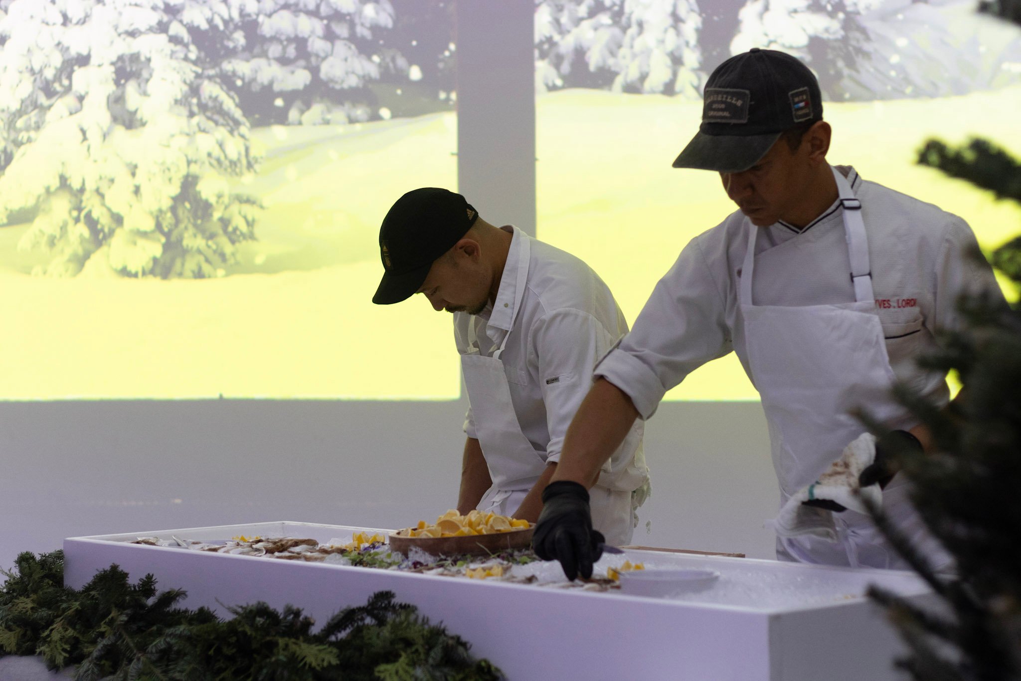 Two chefs in white uniforms prepare garnished seafood at a counter beneath a projected landscape.