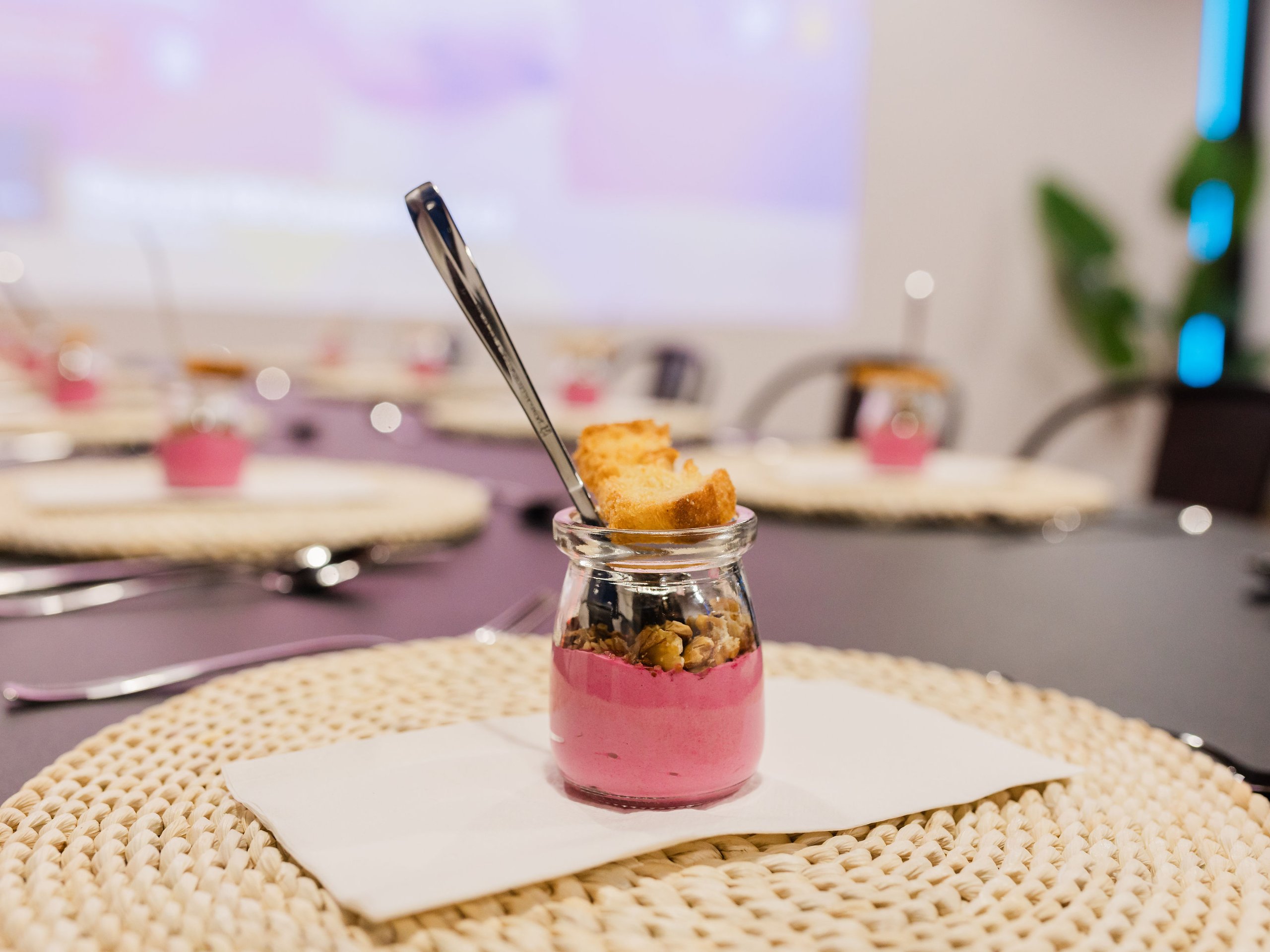 Pink layered dessert in a glass jar topped with nuts and biscuits, with a spoon on a white plate.