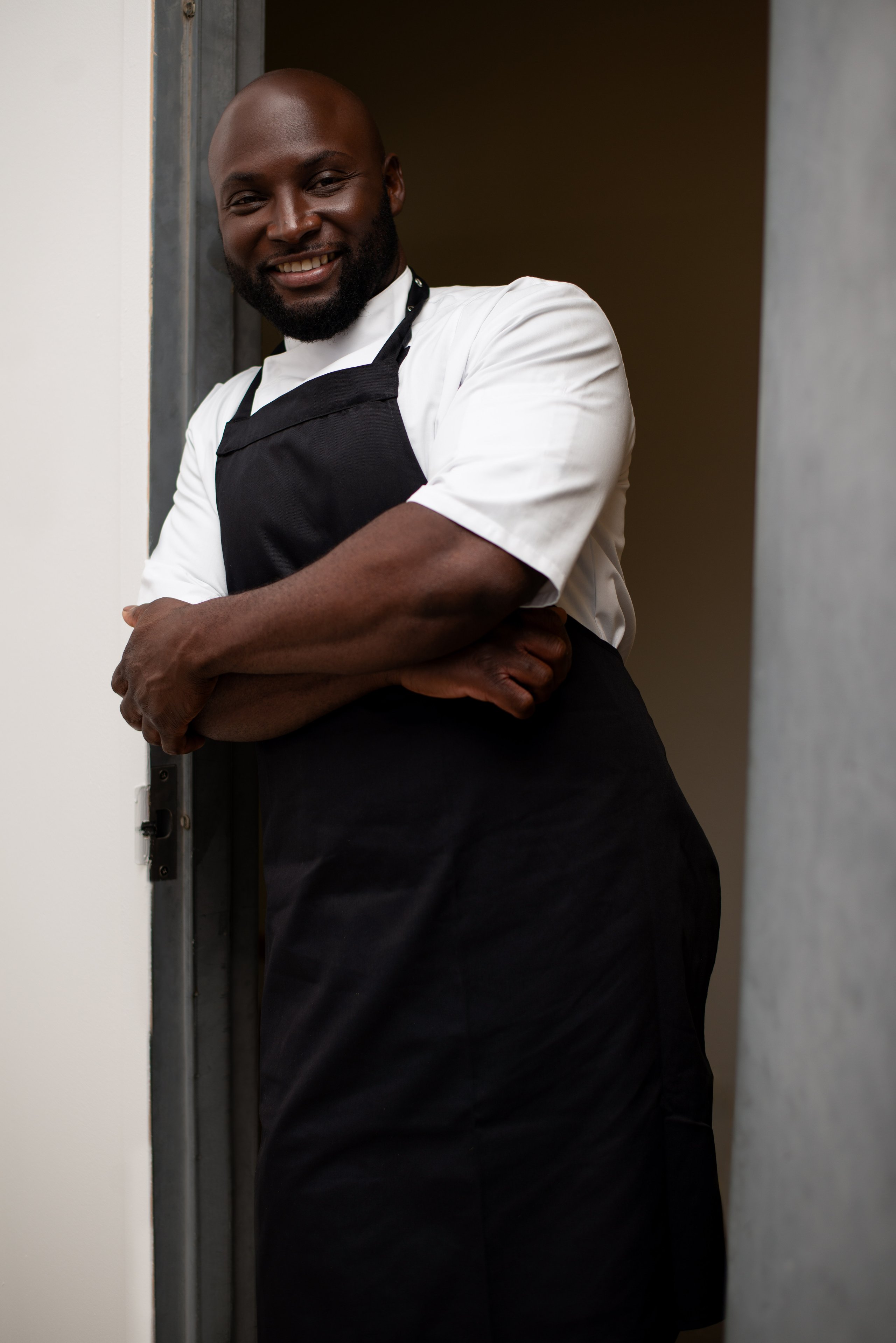 A smiling Black chef with a beard and shaved head standing in a doorway with crossed arms, wearing a black apron over a white jacket.